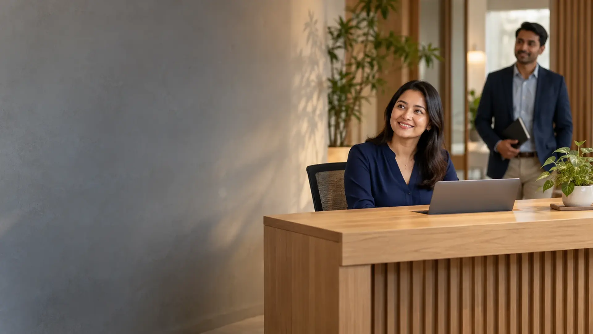 A smiling receptionist at a wooden front desk with a laptop, a colleague holding a notebook behind her
